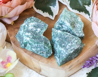 May include: Four rough, green aventurine crystal chunks on a wooden surface. The crystals are a light green color with a speckled appearance.