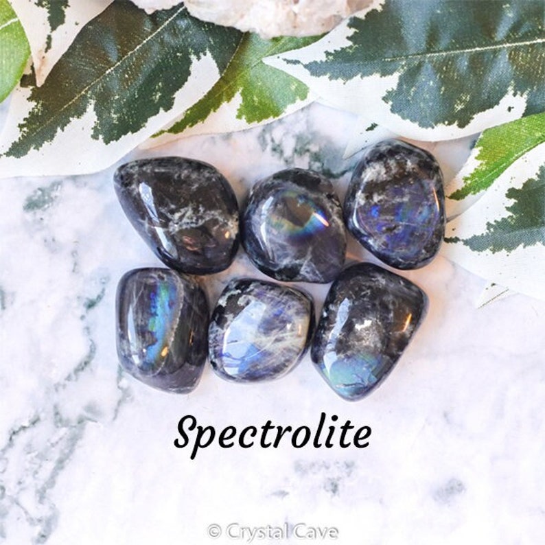 May include: A collection of six smooth, polished spectrolite stones. The stones are dark grey with iridescent blue, green, and purple hues. The stones are arranged on a white marble surface. The text "Spectrolite" is written in black below the stones. The text "Crystal Cave" is written in black below the word "Spectrolite".