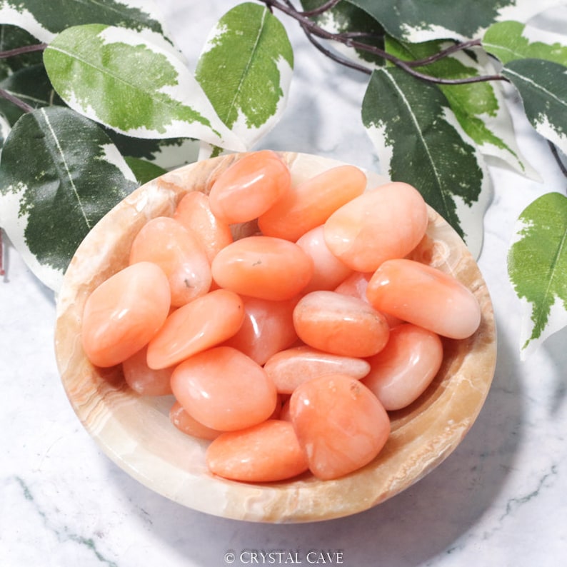 May include: A bowl filled with smooth, orange carnelian tumbled stones. The stones are a light orange color and have a natural, polished finish.