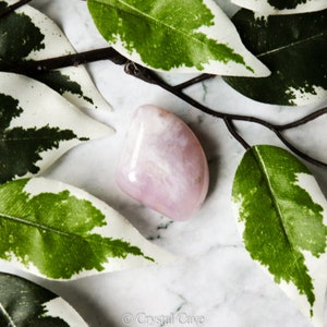 Pode incluir: Um cristal rosa e branco polido e liso com uma forma natural irregular. O cristal est&aacute; apoiado numa superf&iacute;cie de m&aacute;rmore branco com folhas verdes e brancas.