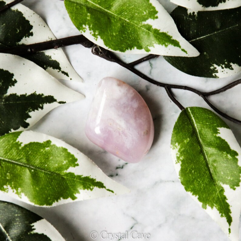 Pode incluir: Um cristal rosa e branco polido e liso com uma forma natural irregular. O cristal est&aacute; apoiado numa superf&iacute;cie de m&aacute;rmore branco com folhas verdes e brancas.