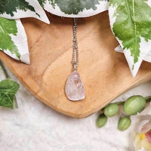 Puede incluir: Un collar de cadena plateada con un colgante de cristal transparente, rosa claro y transl&uacute;cido. El colgante tiene una forma libre y tiene un aspecto natural y en bruto.