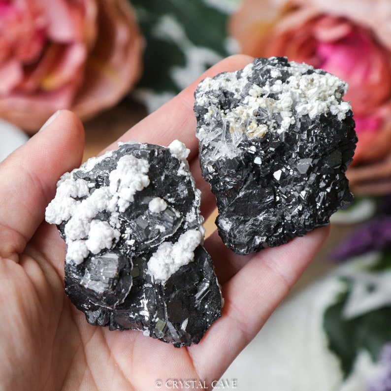 May include: Two black and white mineral specimens with a rough, textured surface. The larger specimen is on the left and the smaller specimen is on the right. Both specimens have white crystals on the surface.