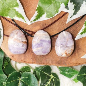 Puede incluir: Tres piedras preciosas ovaladas pulidas, en tonos lavanda, beige y marr&oacute;n, se presentan sobre una superficie de madera. Cada piedra est&aacute; ensartada en un cord&oacute;n de cuero. El fondo incluye hojas verdes y blancas, creando una est&eacute;tica natural.