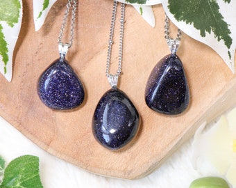 May include: Three silver necklaces with blue gemstone pendants. The pendants are teardrop shaped and have a sparkly, speckled texture. The necklaces are displayed on a wooden surface.