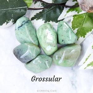 Pode incluir: Uma coleção de seis pedras de granadas grossulares lisas, verdes e translúcidas. As pedras estão dispostas sobre uma superfície branca com folhas verdes no fundo.