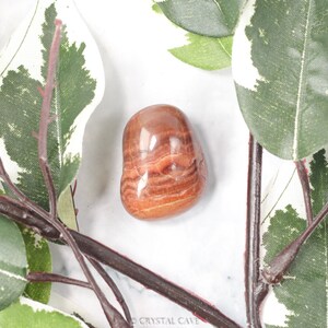 May include: A smooth, polished red and brown banded agate tumbled stone. The stone has a natural, organic shape and is perfect for crystal healing or meditation.