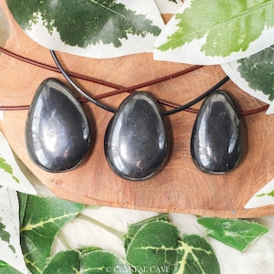 May include: Three black hematite teardrop pendants on a brown leather cord. The pendants are different sizes and are arranged in a row on a wooden surface.