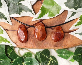 May include: Three orange carnelian stones on a brown leather cord necklace. The stones are smooth and polished, and they are arranged in a row on a wooden surface.