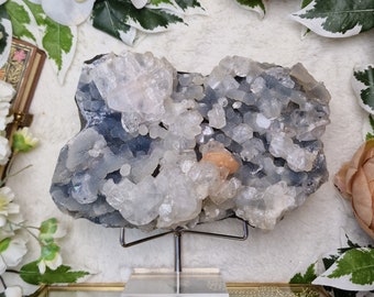 May include: A large, white, crystal cluster with a dark gray base. The cluster is on a metal stand with a clear acrylic base. The crystal cluster is surrounded by white flowers.
