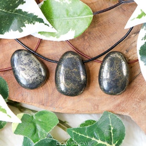 May include: Three polished pyrite pendants on leather cords. The stones have a metallic, speckled appearance with a dark, gunmetal gray base color and gold flecks. The pendants are displayed on a wooden surface, surrounded by green and white foliage.