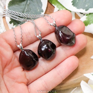 May include: Three silver necklaces with dark red gemstone pendants. The pendants are smooth and have a natural, irregular shape. The necklaces are on a wooden surface with green leaves in the background.