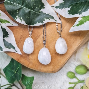 May include: Three white gemstone pendants on silver chains. The pendants are smooth and oval-shaped. The pendants are displayed on a wooden surface with green leaves in the background.