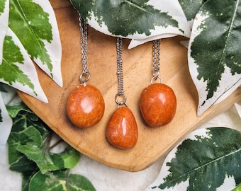 Op de afbeelding: Drie oranje carneol-stenen hangers aan zilveren kettingen. De stenen zijn glad en gepolijst en hangen aan een houten oppervlak.