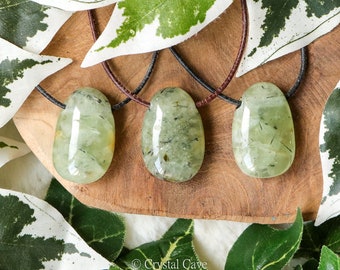 May include: Three prehnite gemstone pendants on brown leather cords. The polished, oval-shaped stones are a translucent light green with dark inclusions. The pendants are displayed on a wooden surface, surrounded by green and white leaves.