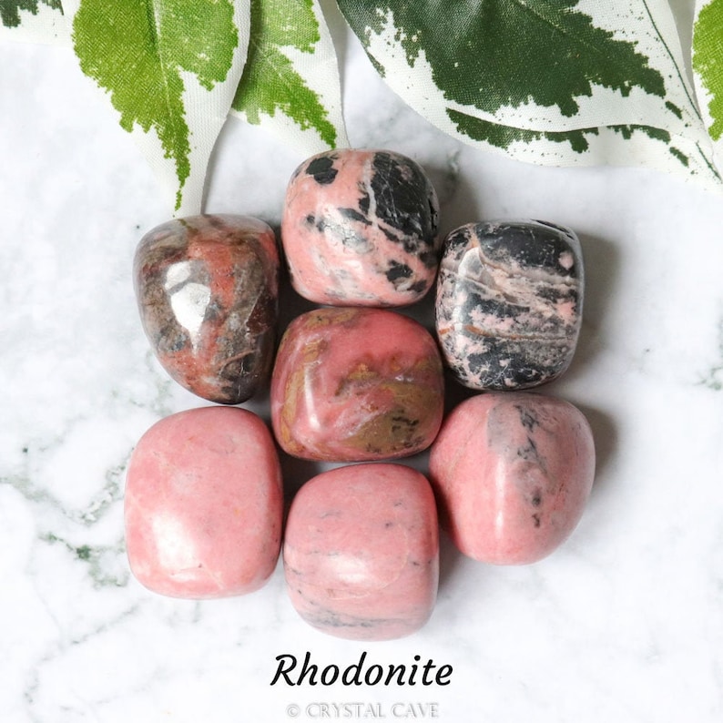 May include: A collection of seven tumbled rhodonite stones. The stones are a mix of pink, black, and brown colors. The stones are arranged in a circle on a white marble surface.