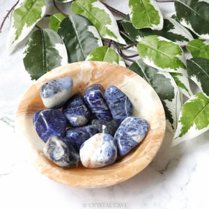 May include: A bowl of blue and white tumbled Sodalite stones. The stones are smooth and polished, and they are arranged in a bowl made of light brown marble.