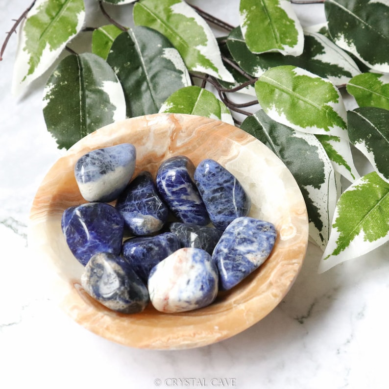 May include: A bowl of blue and white tumbled Sodalite stones. The stones are smooth and polished, and they are arranged in a bowl made of light brown marble.