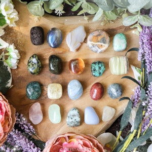 May include: A collection of 20 tumbled stones in various colors and textures, including green, blue, red, orange, yellow, and white. The stones are arranged on a wooden surface.