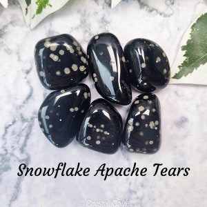 Op de afbeelding: Zes gladde, zwarte sneeuwvlok-obsidiaan-tumbled stones met witte insluitsels. De stenen zijn in een cirkel op een wit marmeren oppervlak gerangschikt.