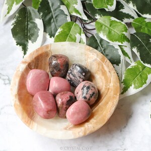 May include: A collection of seven pink and black tumbled stones in a light brown bowl. The stones are smooth and polished, and they have a variety of shapes and sizes.