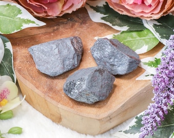 May include: Three rough, gray hematite stones on a wooden surface. The stones are irregularly shaped and have a metallic sheen.
