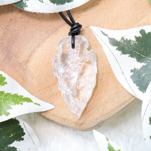 May include: A clear quartz arrowhead pendant on a black leather cord. The arrowhead is a natural crystal with a rough, irregular shape. The pendant is hanging on a wooden surface with green leaves in the background.