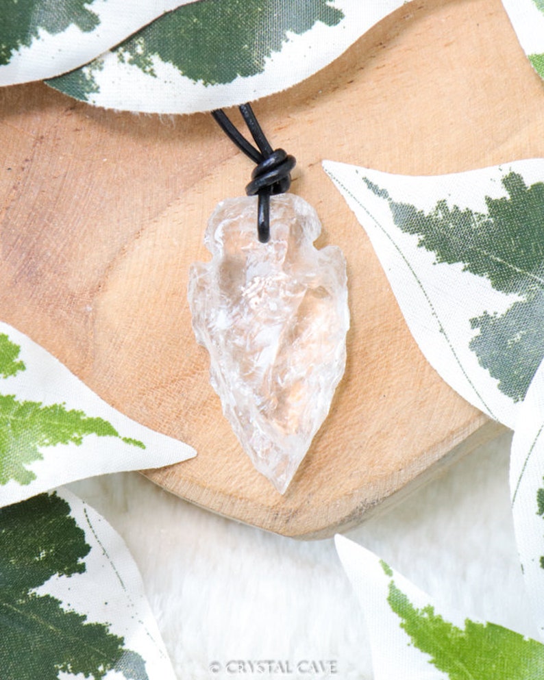 May include: A clear quartz arrowhead pendant on a black leather cord. The arrowhead is a natural crystal with a rough, irregular shape. The pendant is hanging on a wooden surface with green leaves in the background.