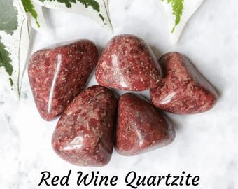 May include: Five tumbled red wine quartzite stones. The stones are a deep red color with white and gray veins. The stones are arranged in a cluster on a white marble surface.
