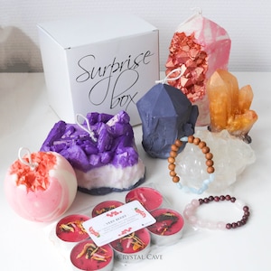 Pode incluir: Uma caixa branca com o texto "Surprise Box" nela. A caixa é cercada por várias velas em forma de cristal, incluindo roxo, rosa, laranja e azul. Há também duas pulseiras e um conjunto de velas de chá com o texto "Very Berry" na embalagem.