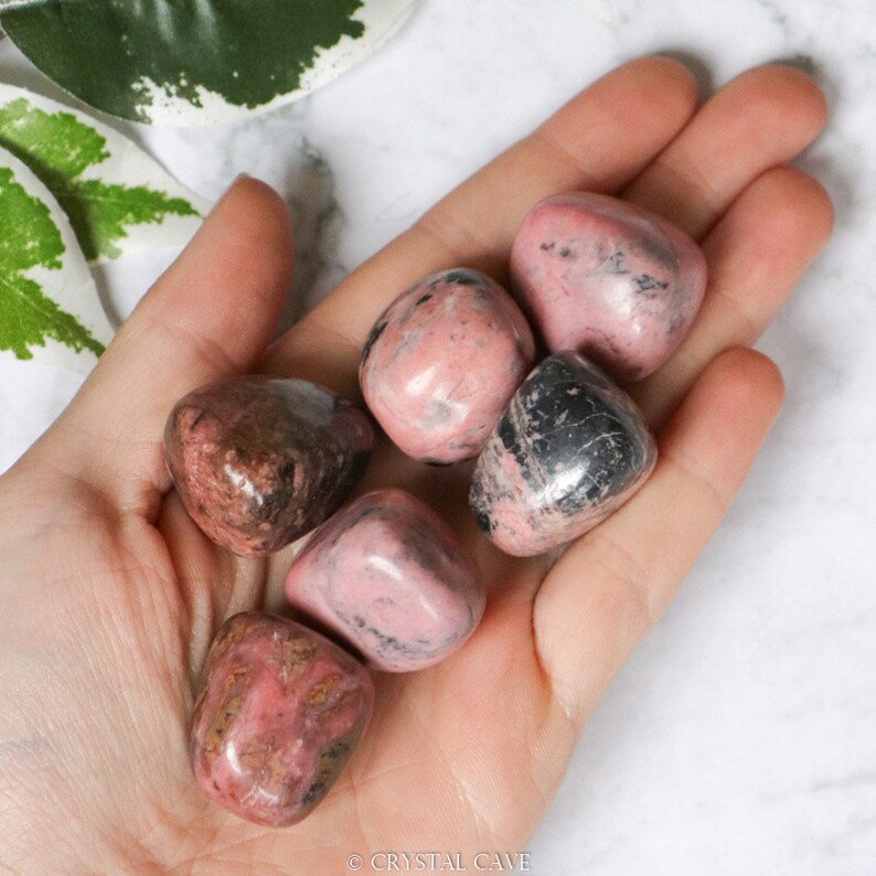 May include: Six tumbled pink and black rhodonite stones in a hand. The stones are smooth and have a natural, polished finish.