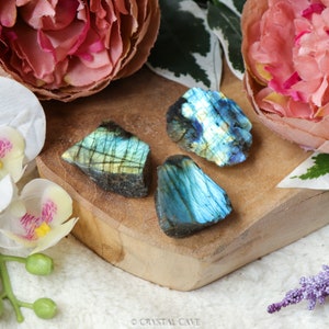 Op de afbeelding: Drie ruwe labradorietstenen met iriserende blauwe, groene en gele flitsen worden getoond op een houten plank. De stenen zijn omringd door roze bloemen en witte orchideeën, wat een natuurlijk en decoratief arrangement creëert.