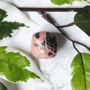 May include: A pink and black tumbled rhodonite stone. The stone has a smooth, polished surface and is a popular choice for crystal healing.