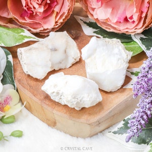 May include: Three white, opaque, rough-textured, natural, raw, crystal clusters on a wooden surface. The crystals are white with a slight off-white hue.