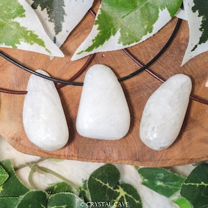 May include: Three white, smooth, teardrop-shaped stones on brown leather cords. The stones are hanging from a wooden surface with green leaves in the background.