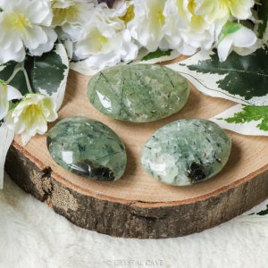 May include: Three oval prehnite stones with green and black inclusions rest on a wooden slice. White flowers and green leaves with white edges surround the stones, creating a natural aesthetic. The stones are smooth and polished.