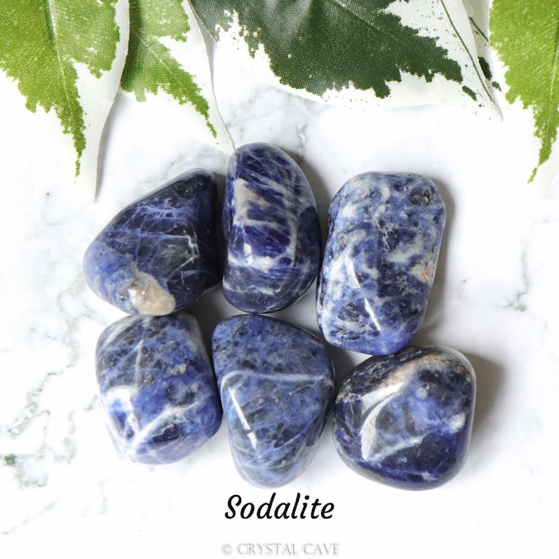 May include: Six tumbled sodalite stones, each with a unique blue and white pattern, on a white marble surface. The stones are smooth and polished, with a natural, earthy feel. The text "Sodalite" is printed below the stones, along with the copyright symbol and "CRYSTAL CAVE".