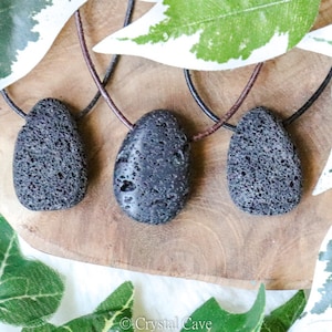 Peut inclure: Trois pendentifs en pierre de lave noire sur des cordons en cuir marron. Les pierres sont lisses et ont une texture rugueuse et poreuse. Les pendentifs sont présentés sur une surface en bois avec des feuilles vertes en arrière-plan.