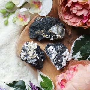 May include: Three black, gray, and white mineral specimens with a rough, natural texture. The specimens are arranged on a wooden surface.