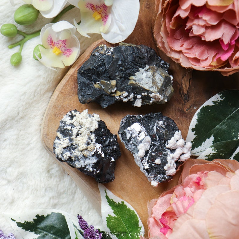 May include: Three black, gray, and white mineral specimens with a rough, natural texture. The specimens are arranged on a wooden surface.