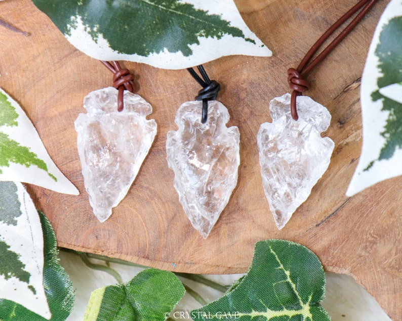 May include: Three clear quartz arrowhead pendants on brown and black leather cords. The arrowheads are polished and have a translucent appearance. The cords are tied in a knot at the top of each pendant.