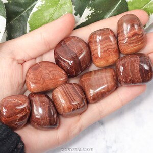 May include: A collection of eight smooth, polished red and brown banded jasper tumbled stones. The stones are various sizes and shapes, with a glossy finish.