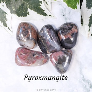 Puede incluir: Cinco piedras pulidas de Pyroxmangita, un mineral gris oscuro, rosa y blanco. Las piedras son lisas y pulidas, con una forma natural y orgánica. El texto "Pyroxmangite" está escrito en negro debajo de las piedras.