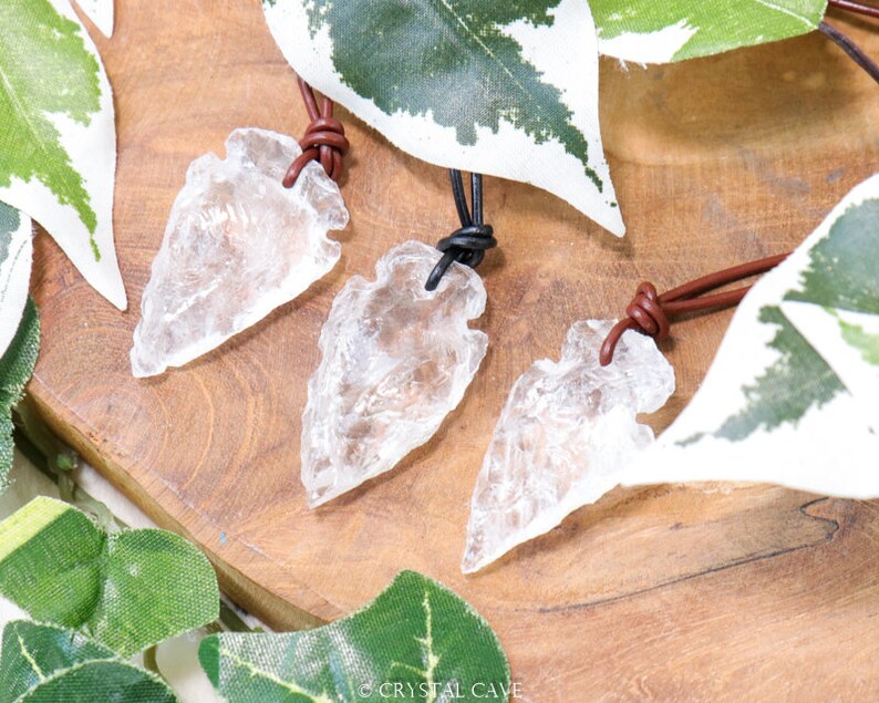 May include: Three clear quartz arrowhead pendants on brown leather cords. The arrowheads are on a wooden surface with green leaves.