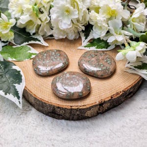 May include: Three green and brown jasper palm stones on a wooden surface. The stones are smooth and polished.