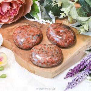 Könnte beinhalten: Drei glatte, polierte rote und weiße Jaspis-Steine, auch bekannt als roter Aventurin, auf einer Holzoberfläche. Die Steine sind oval geformt und haben ein natürliches, organisches Aussehen.