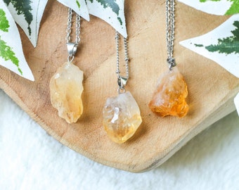 May include: Three gold citrine crystal necklaces on a wooden surface. Each necklace features a raw, natural crystal pendant and a silver chain.