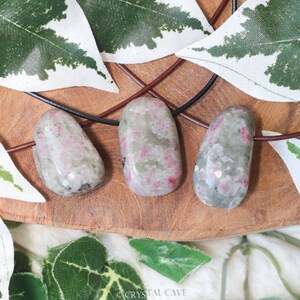 May include: Three pink and green gemstone pendants on a black leather cord. The pendants are smooth and have a natural, organic shape. They are displayed on a wooden surface with green leaves.