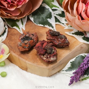 May include: Three small, rough, red and brown, natural, mineral specimens with green crystals embedded in the rock. The specimens are on a wooden surface.