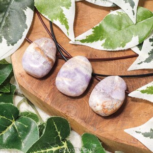 Puede incluir: Tres piedras pulidas de forma ovalada, en tonos lavanda, beige y marr&oacute;n, ensartadas en cordones negros. Se exhiben sobre una superficie de madera, rodeadas de acentos de hojas verdes y blancas. La imagen resalta las texturas y colores naturales de las piedras.
